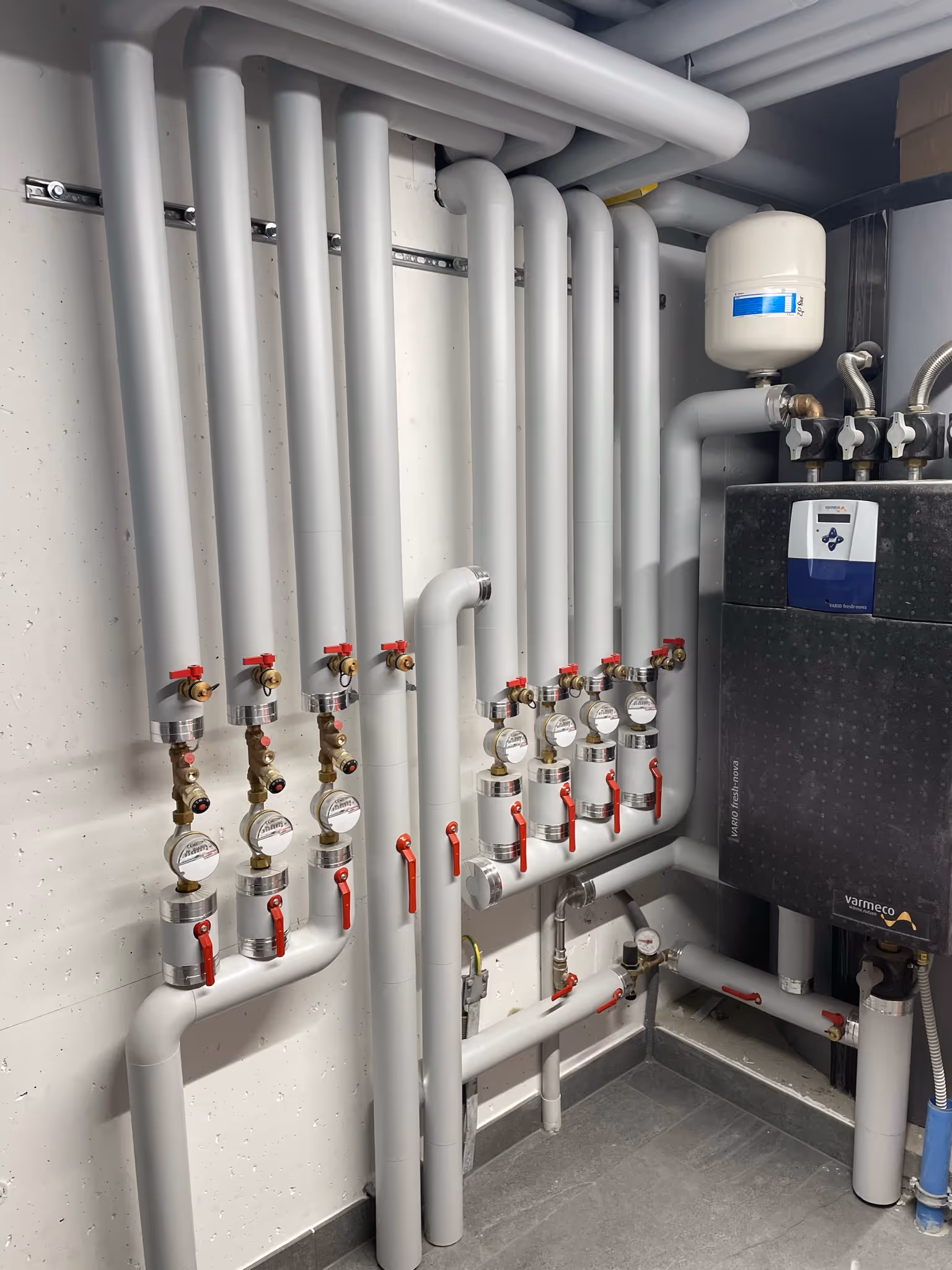Mehrere graue Heizungsrohre mit roten Ventilgriffen, Wasserzählern und einem weißen Ausdehnungsgefäß an der Wand in einem Technikraum.