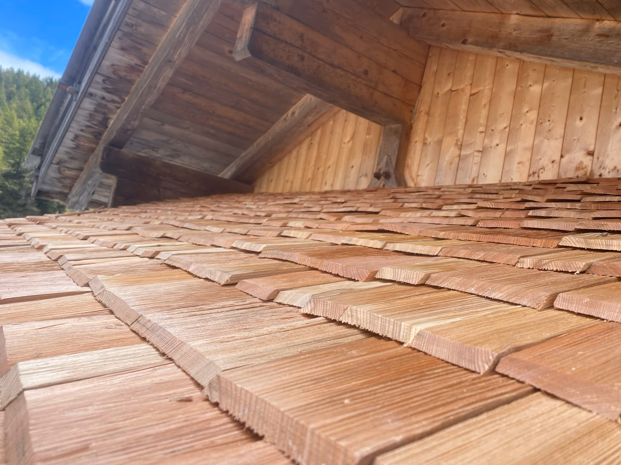 Nahaufnahme eines mit Holzschindeln gedeckten Dachs vor einer Holzwand unter blauem Himmel.