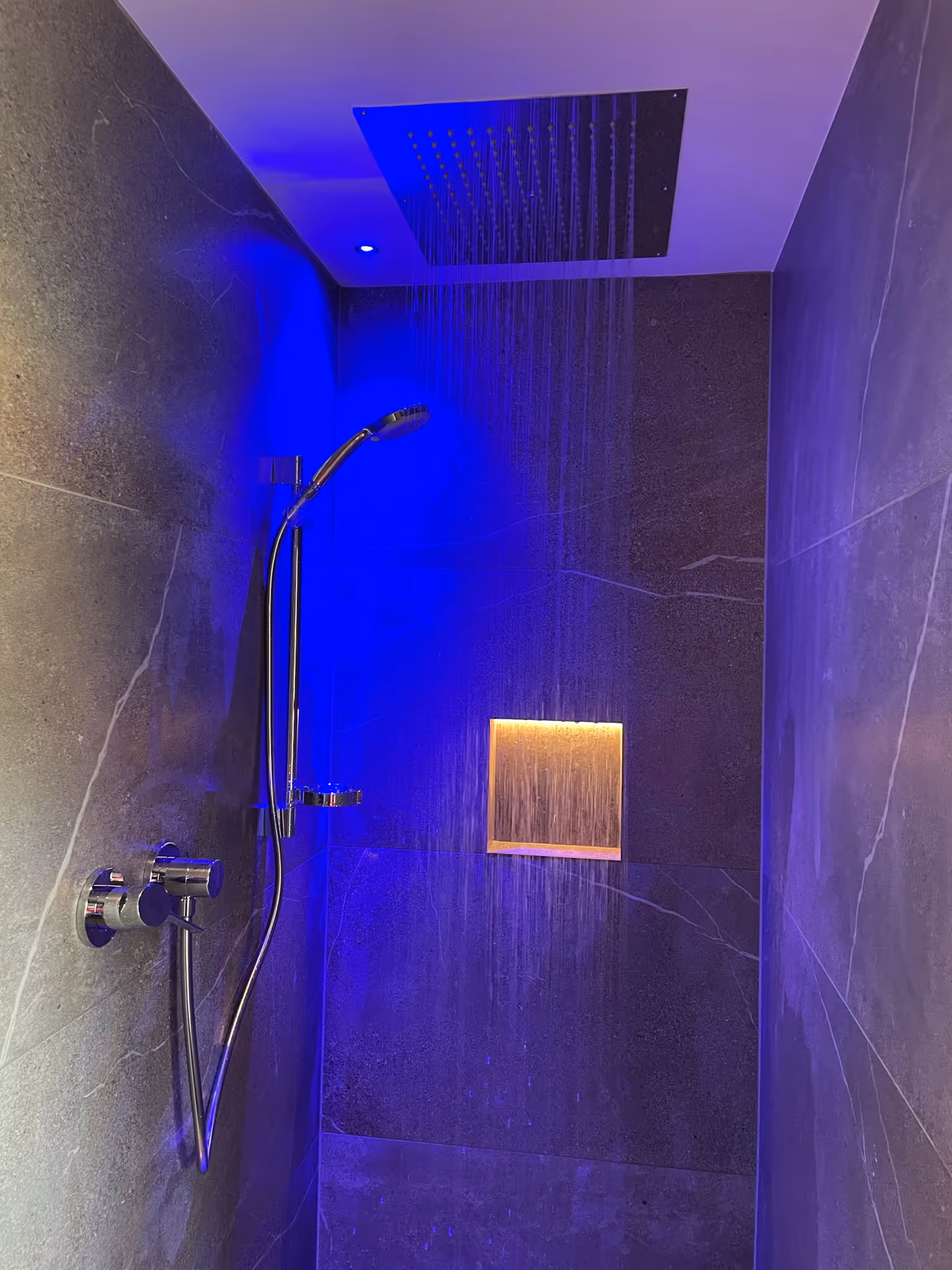 Moderner Duschbereich mit grauen Fliesen, Regenbrause an der Decke und Handbrause, beleuchtet mit blauem Licht.