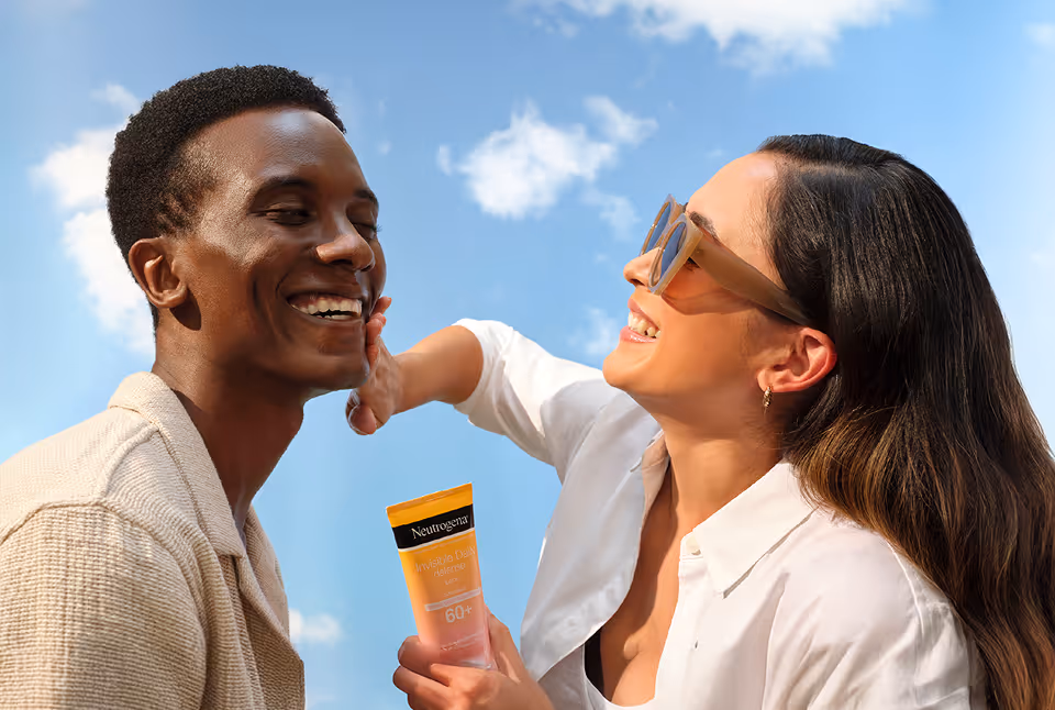 Woman wearing sunglasses applying Neutrogena sunscreen to smiling man's face under a blue sky with clouds.