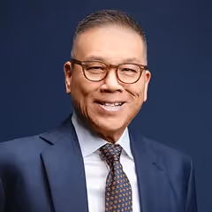 Smiling middle-aged man wearing glasses, a dark suit, white shirt, and patterned tie against a blue background.
