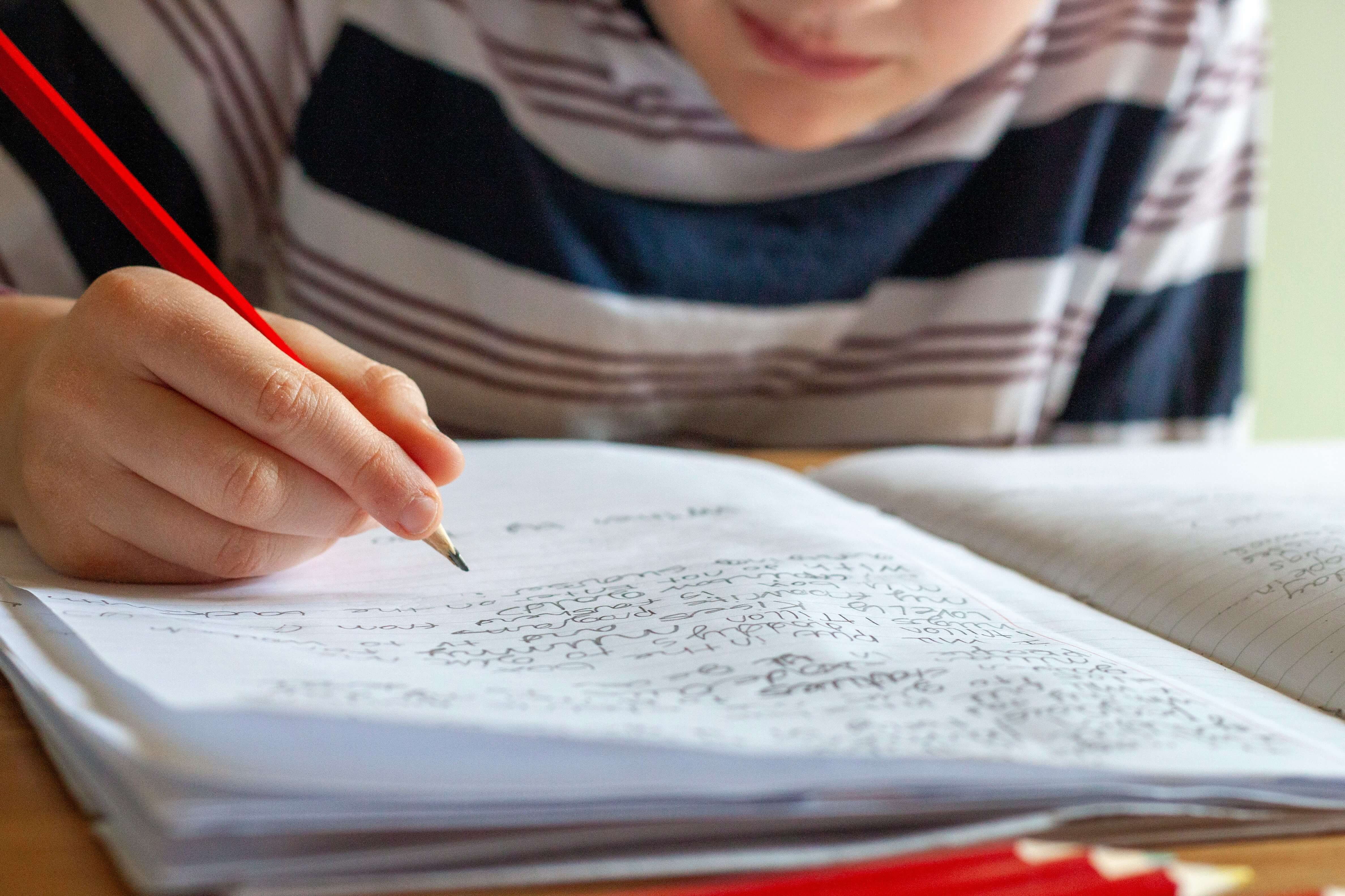 Child writing with a red pencil in a notebook with handwritten notes.