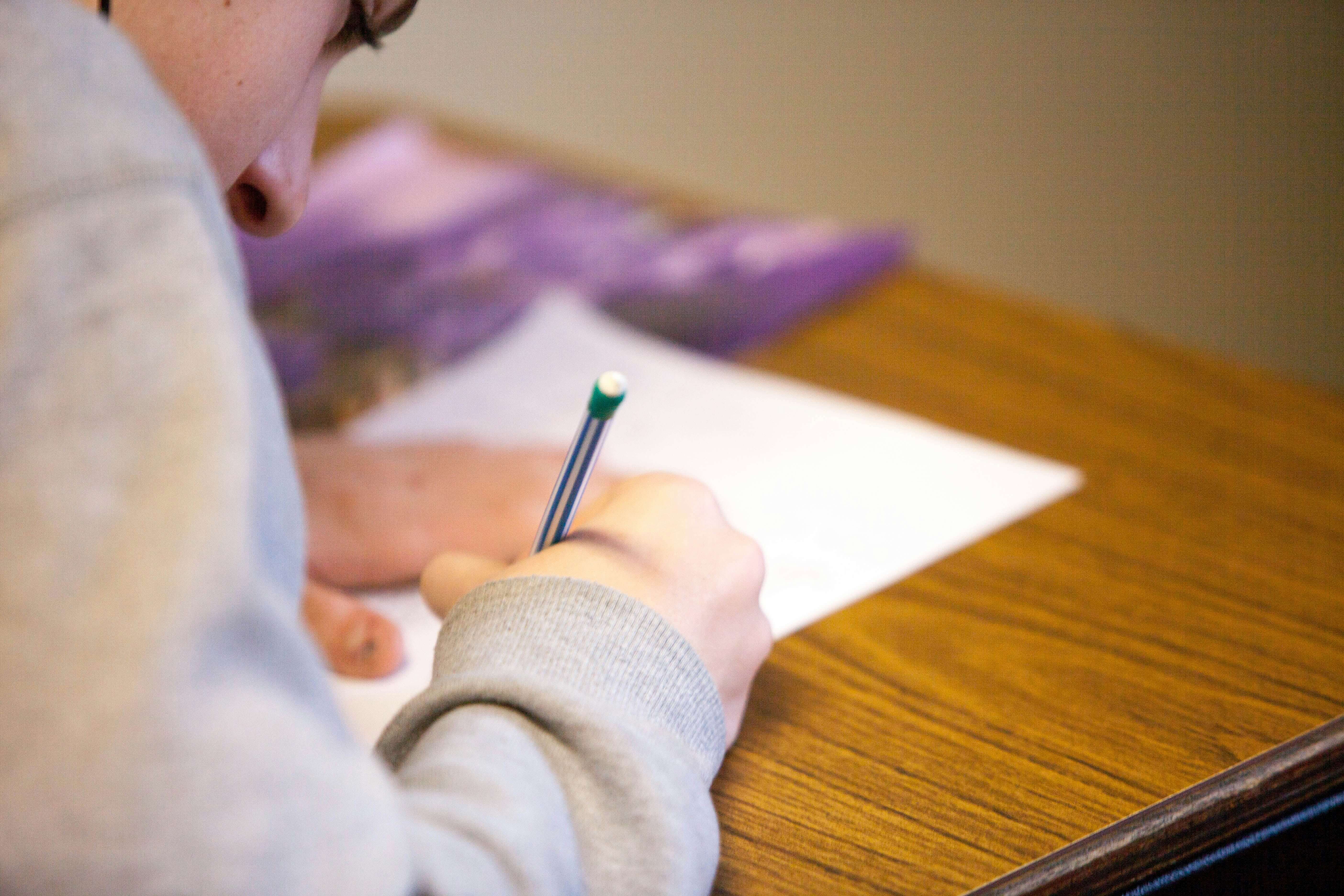 Person writing on paper with a pencil on a wooden desk.