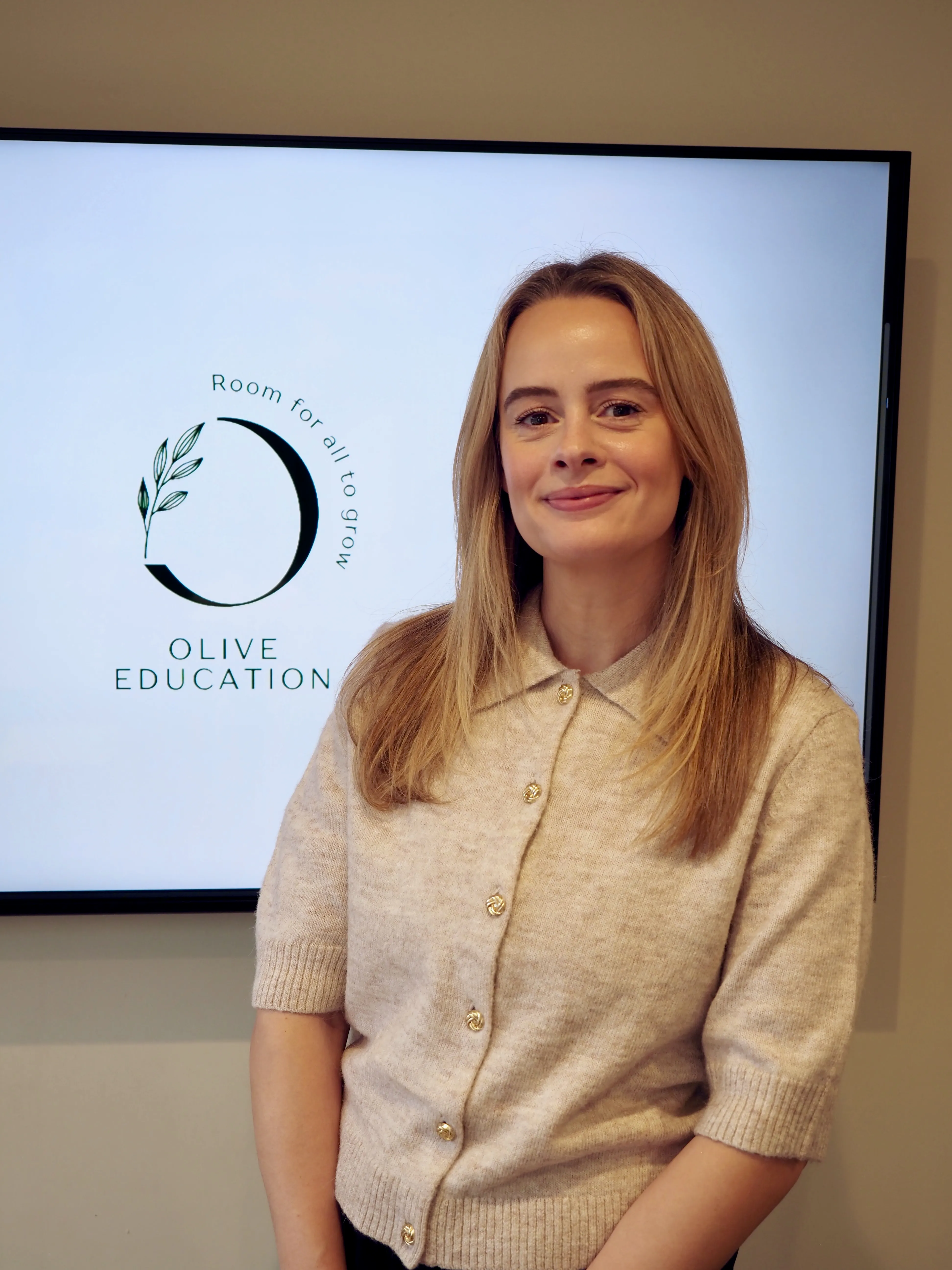 Sarah, the owner, Smiling woman with long blonde hair wearing a beige knit cardigan standing beside a screen displaying Olive Education logo and slogan.