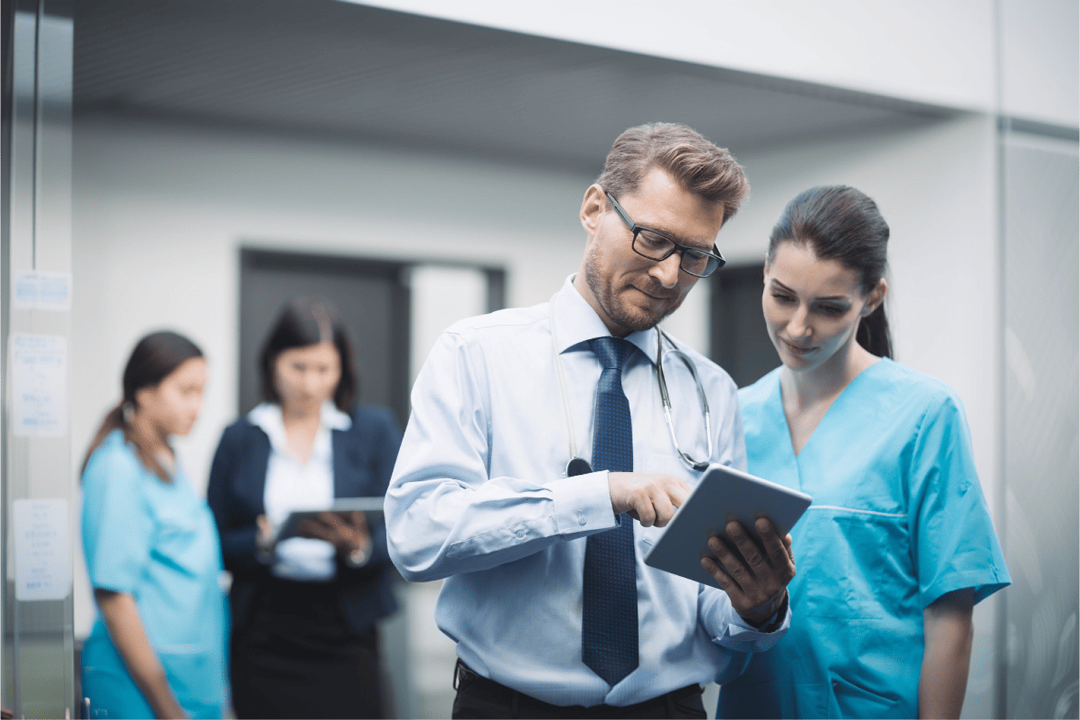  male doctor wearing a stethoscope, dress shirt, and tie reviewing patient information on a digital tablet with a female nurse in teal scrubs, while two other healthcare staff members are visible in the blurred background of a hospital corridor