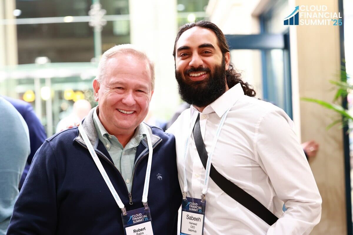 Two men smiling at a conference, one wearing a dark jacket and the other a white shirt with a black strap across the chest.