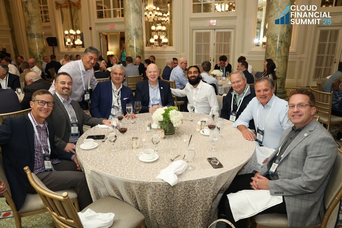 Group of men sitting around a round table with white tablecloth, enjoying drinks and smiling at a Cloud Financial Summit event in an elegant room.