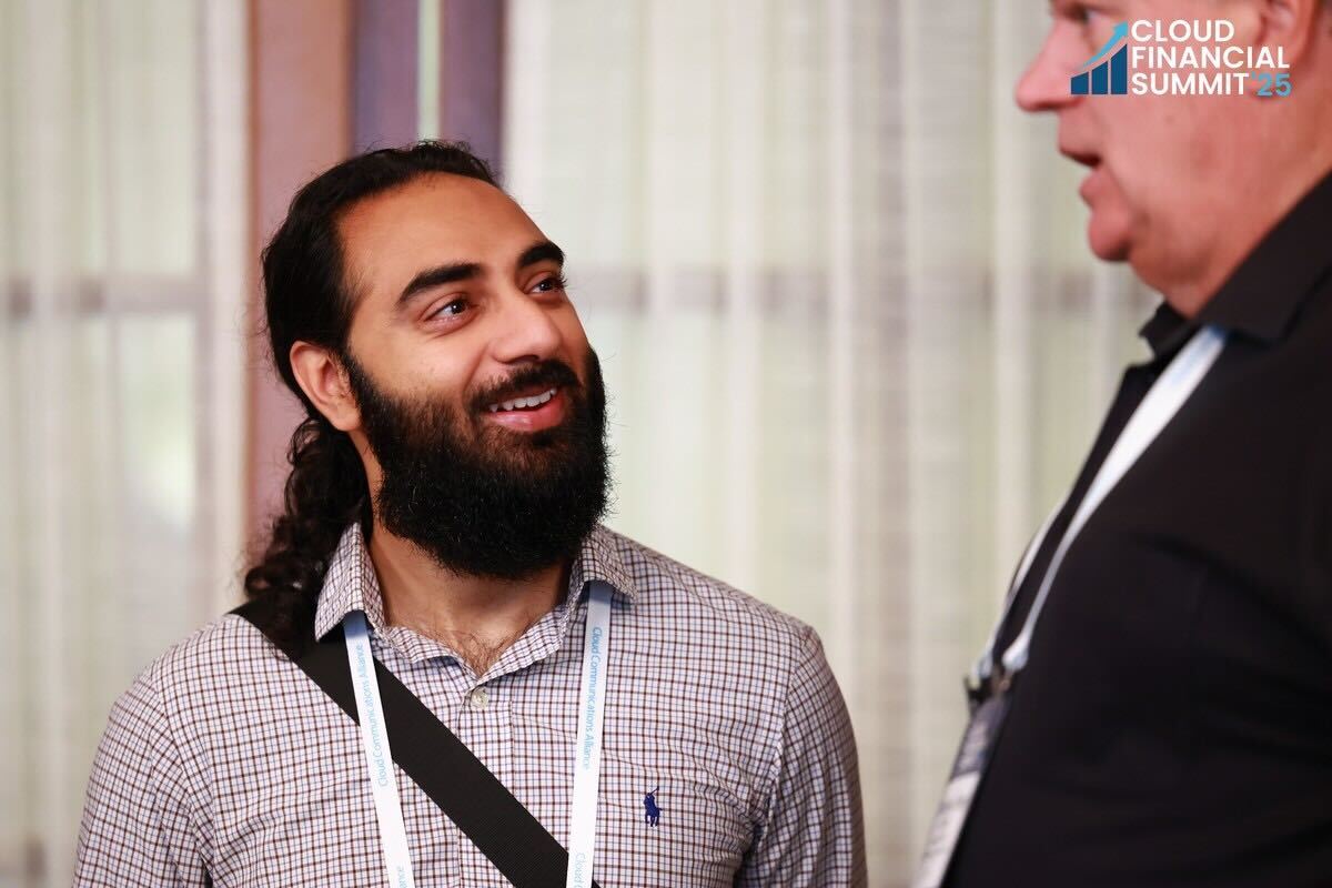 Smiling man with beard and ponytail wearing a checked shirt and lanyard, talking to another person at Cloud Financial Summit.