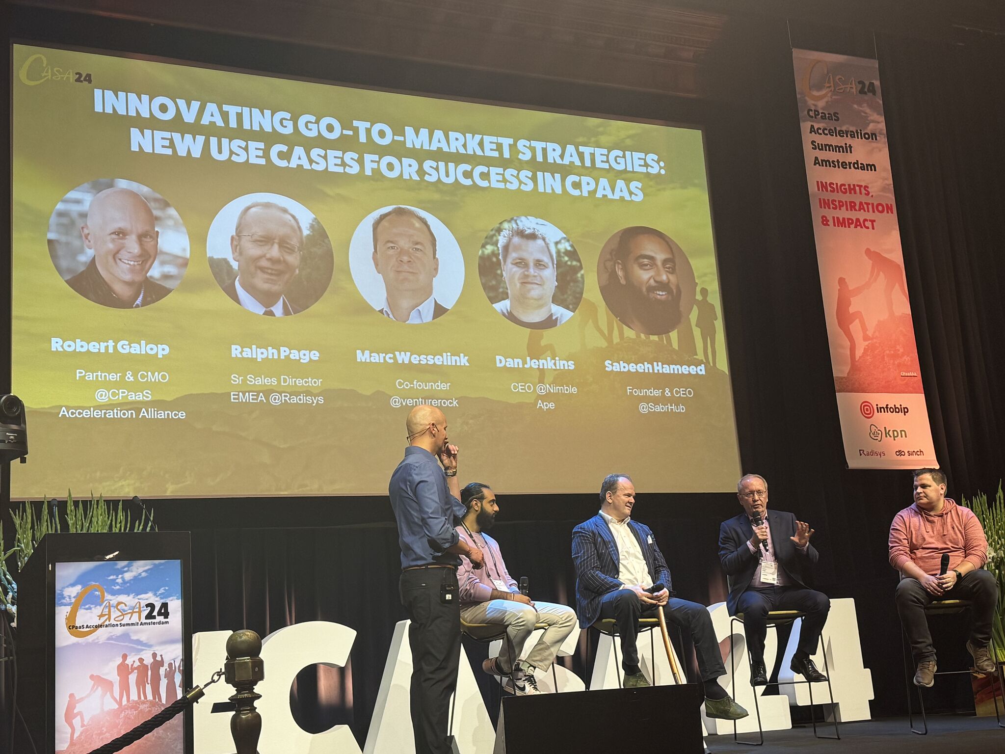 Panel discussion at CPaaS Acceleration Summit Amsterdam with five speakers seated on stage and a presentation slide showing headshots and titles of Robert Galop, Ralph Page, Marc Wessetlink, Dan Jenkins, and Sabeeh Hameed.