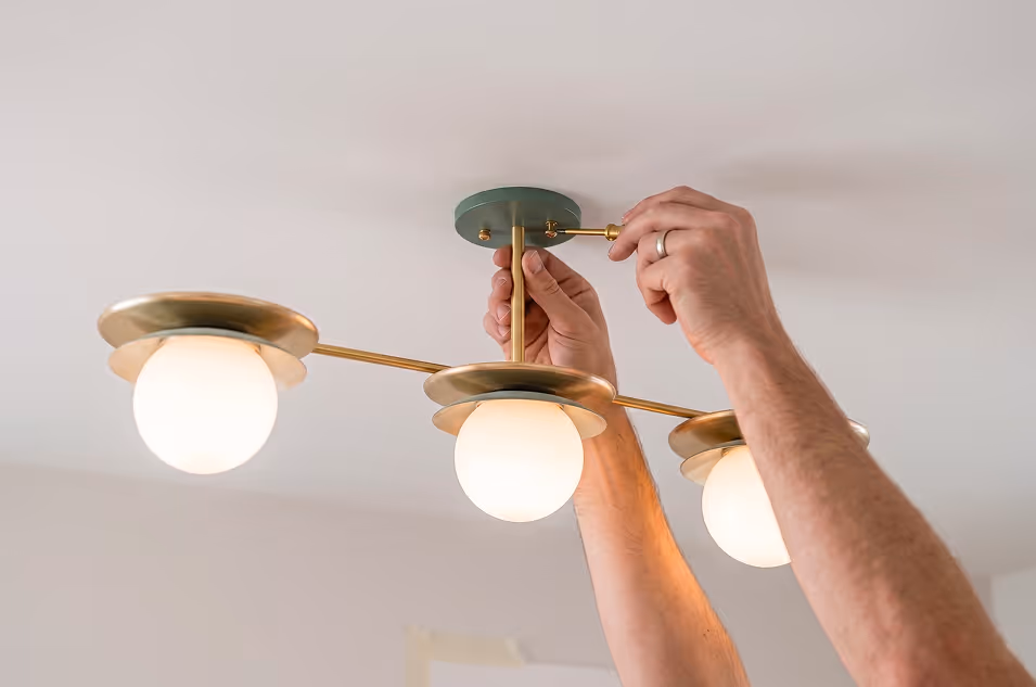 Hands installing a modern brass ceiling light fixture with three glowing round bulbs.