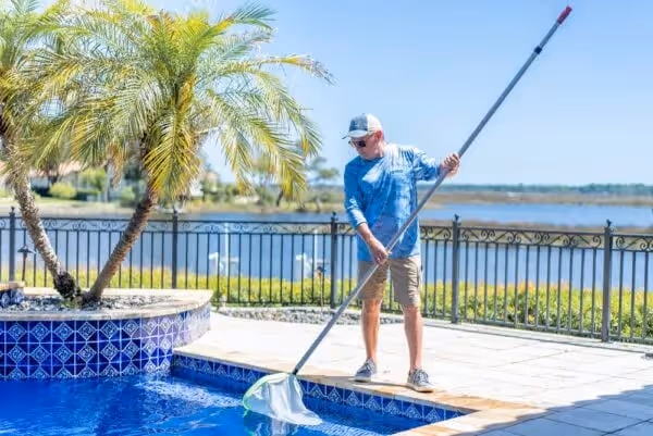 Man in blue shirt and cap cleaning pool with a long-handled net beside palm trees and a lake in the background.