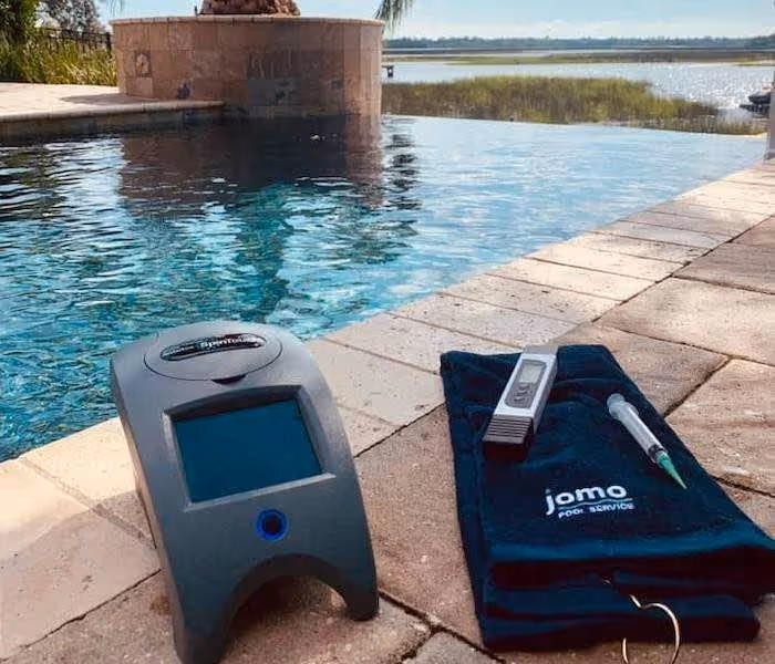 Poolside water testing equipment including a digital meter on a pool deck with a blue towel labeled 'jomo pool service' and a syringe.