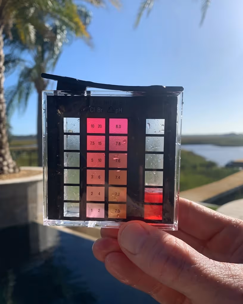 Hand holding a water testing color comparator with pH and chlorine levels against an outdoor background with palm trees and a pool.