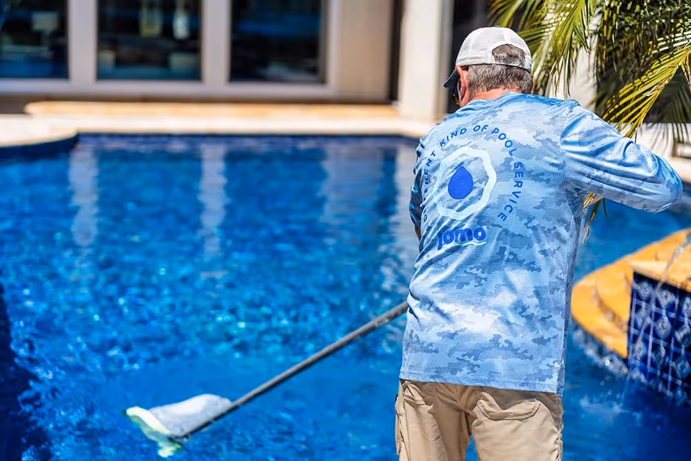 Person cleaning a backyard swimming pool with a long-handled net during sunny day.