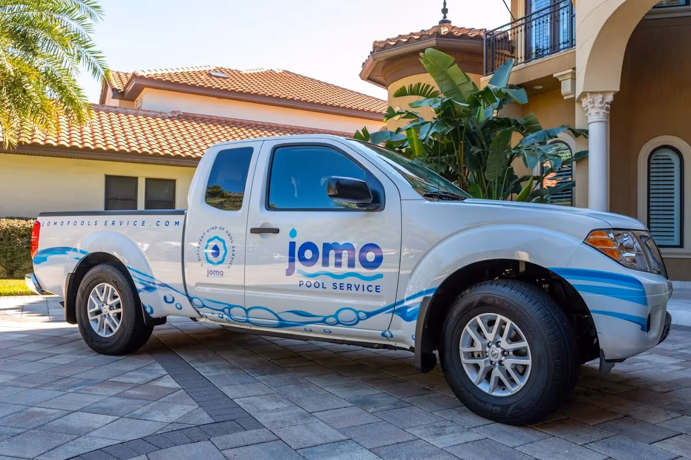 White pickup truck with Jomo Pool Service branding and blue water-themed decals parked on a tiled driveway in front of a house.
