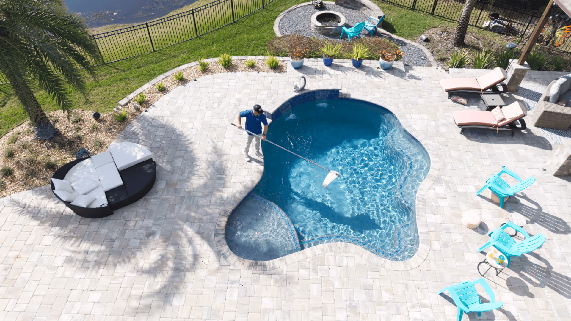 Person cleaning a small kidney-shaped pool with a net skimmer on a sunny patio surrounded by lounge chairs and plants.