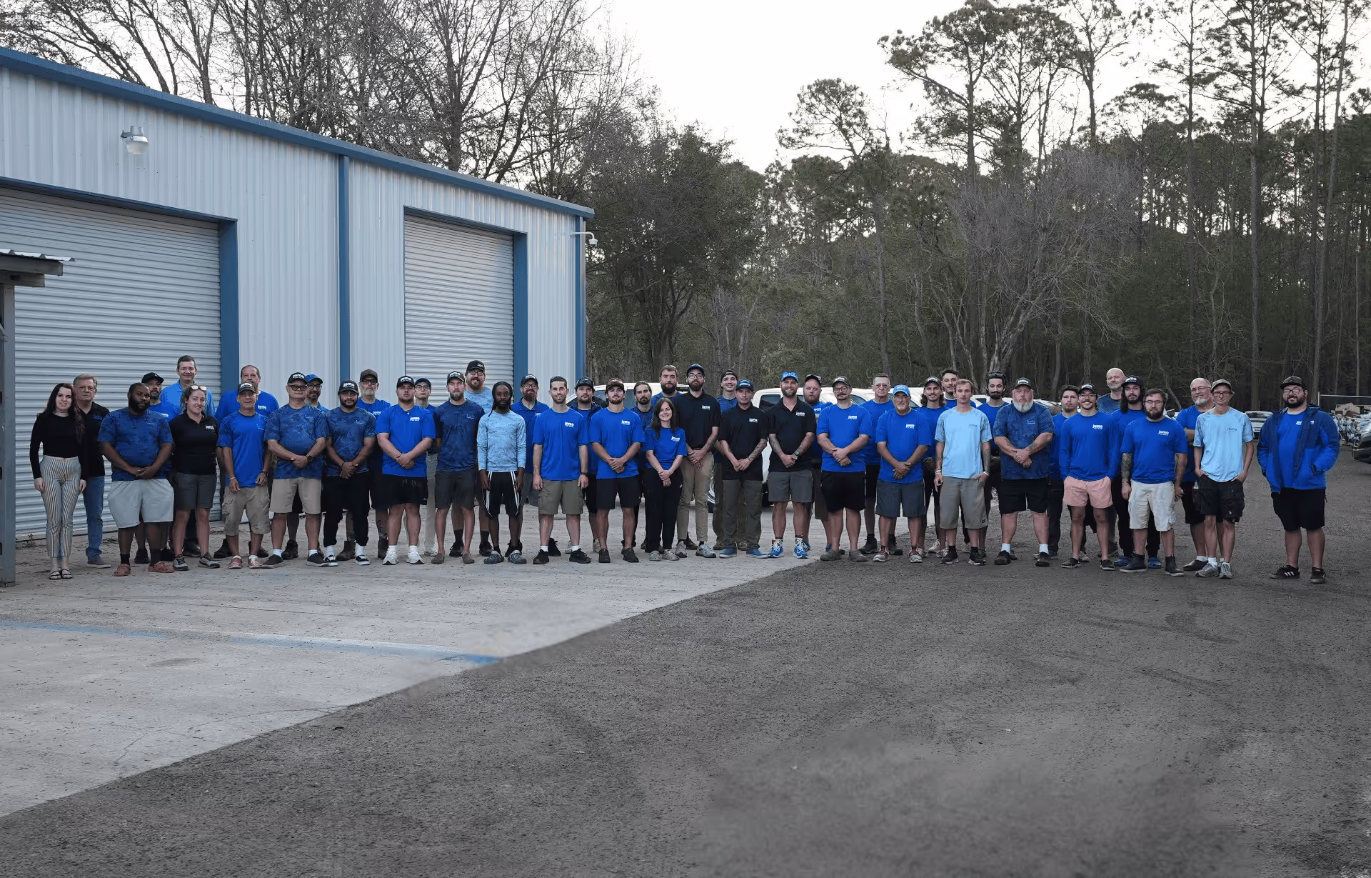 Large group of men and women standing in front of a building with two large closed garage doors, many wearing blue shirts and caps.
