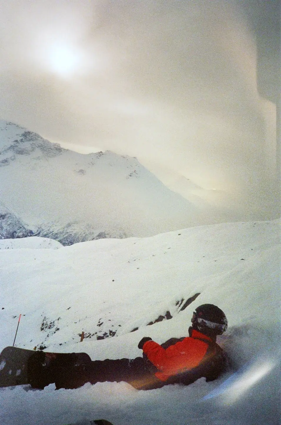 Person in a red jacket and black helmet lying on snow-covered slope with mountains in the background.