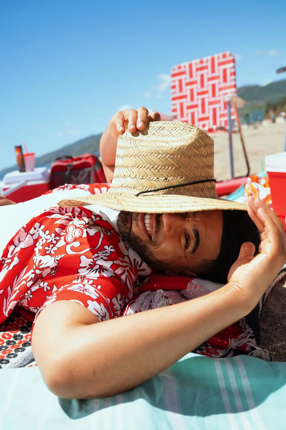 Smiling man wearing a red floral shirt lying on a beach towel while holding a straw hat over his face on a sunny day.