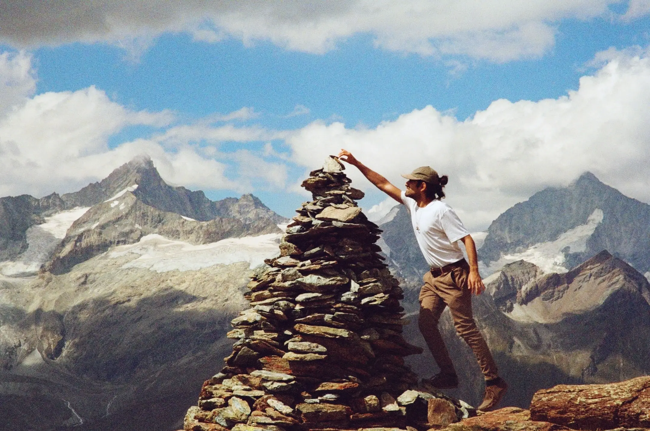 Hiker in a white t-shirt and cap placing a stone on a large stacked rock cairn with snow-capped mountains in the background.