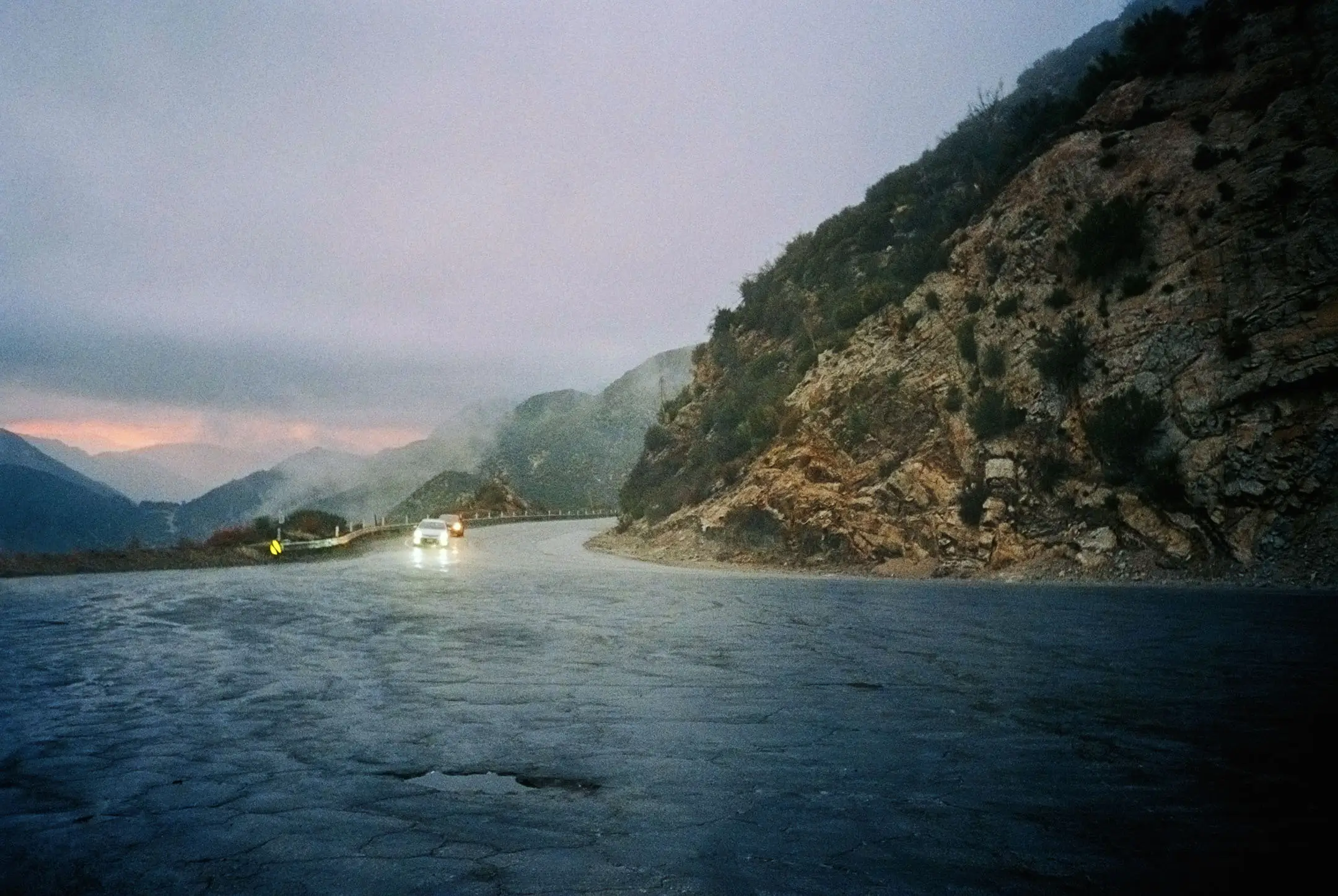 Curved mountain road at dusk with two cars driving towards the camera and rocky hills on the right.