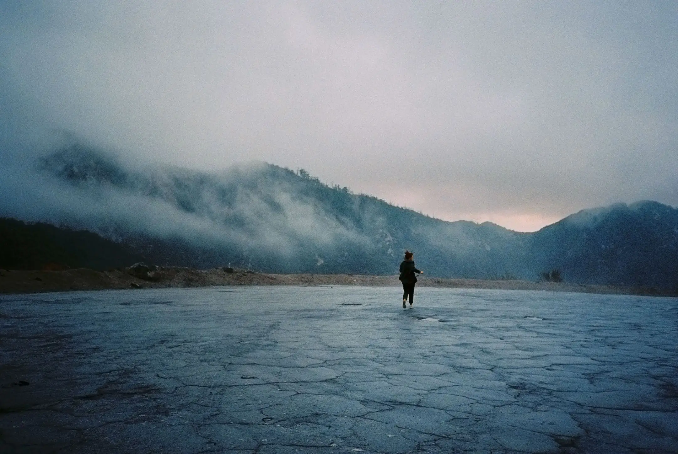 Person walking alone on a cracked, barren landscape with misty mountains in the background under a cloudy sky.