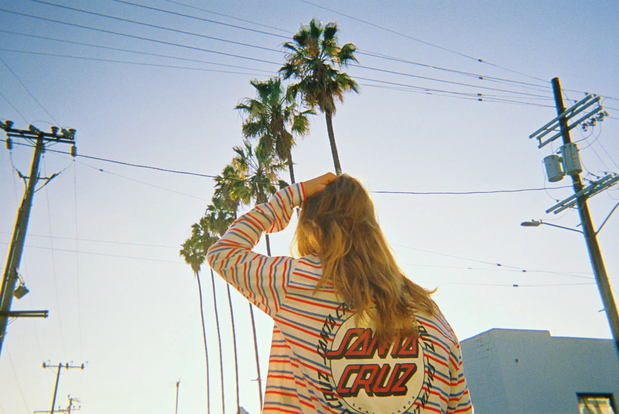 Person with long hair wearing a striped Santa Cruz shirt standing outdoors under palm trees and power lines at sunset.