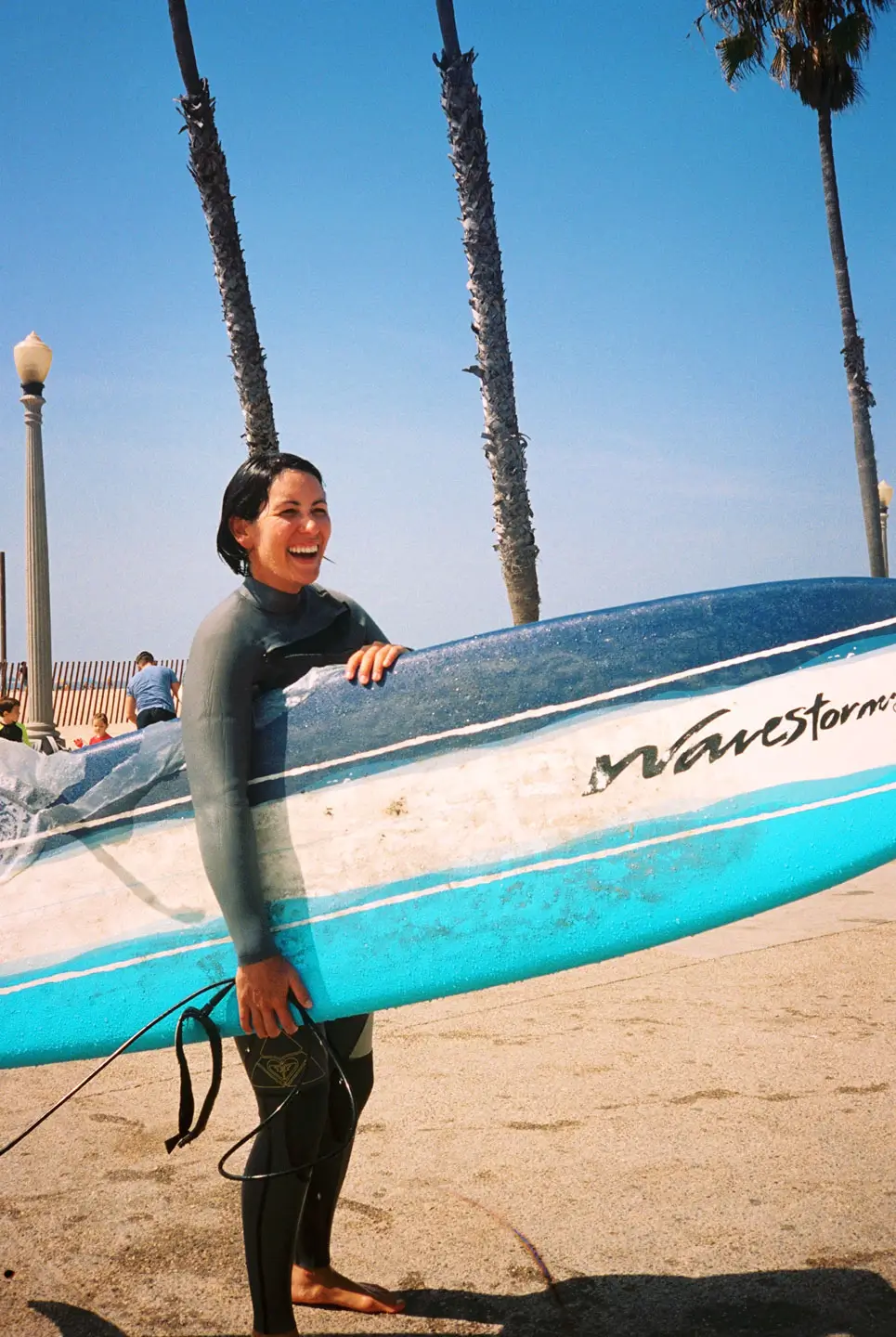 Smiling woman in a wetsuit holding a blue and white surfboard on a sunny beach with palm trees in the background.