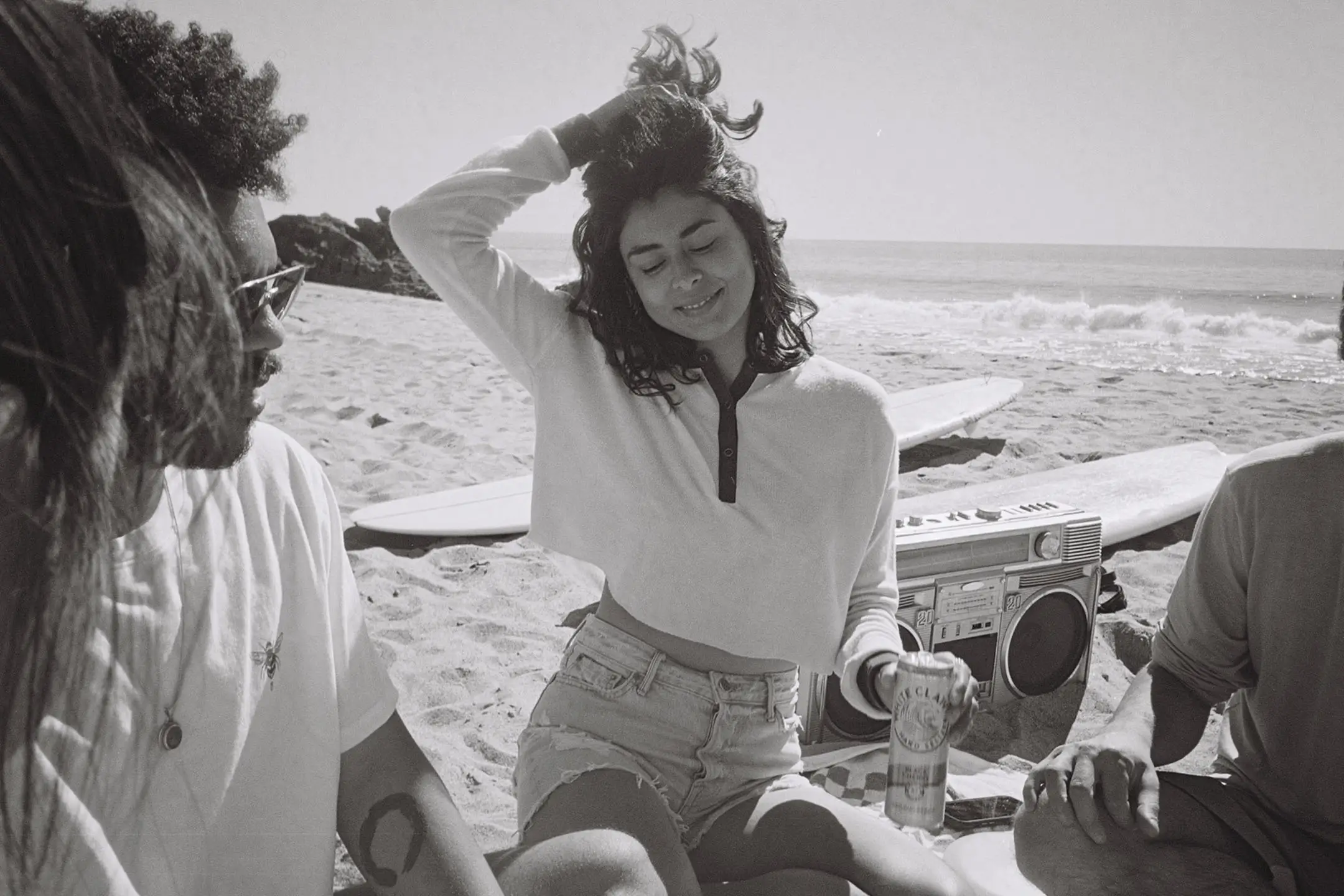 Group of friends sitting on a beach with surfboards and a boombox, one woman smiling and holding a can of White Claw.