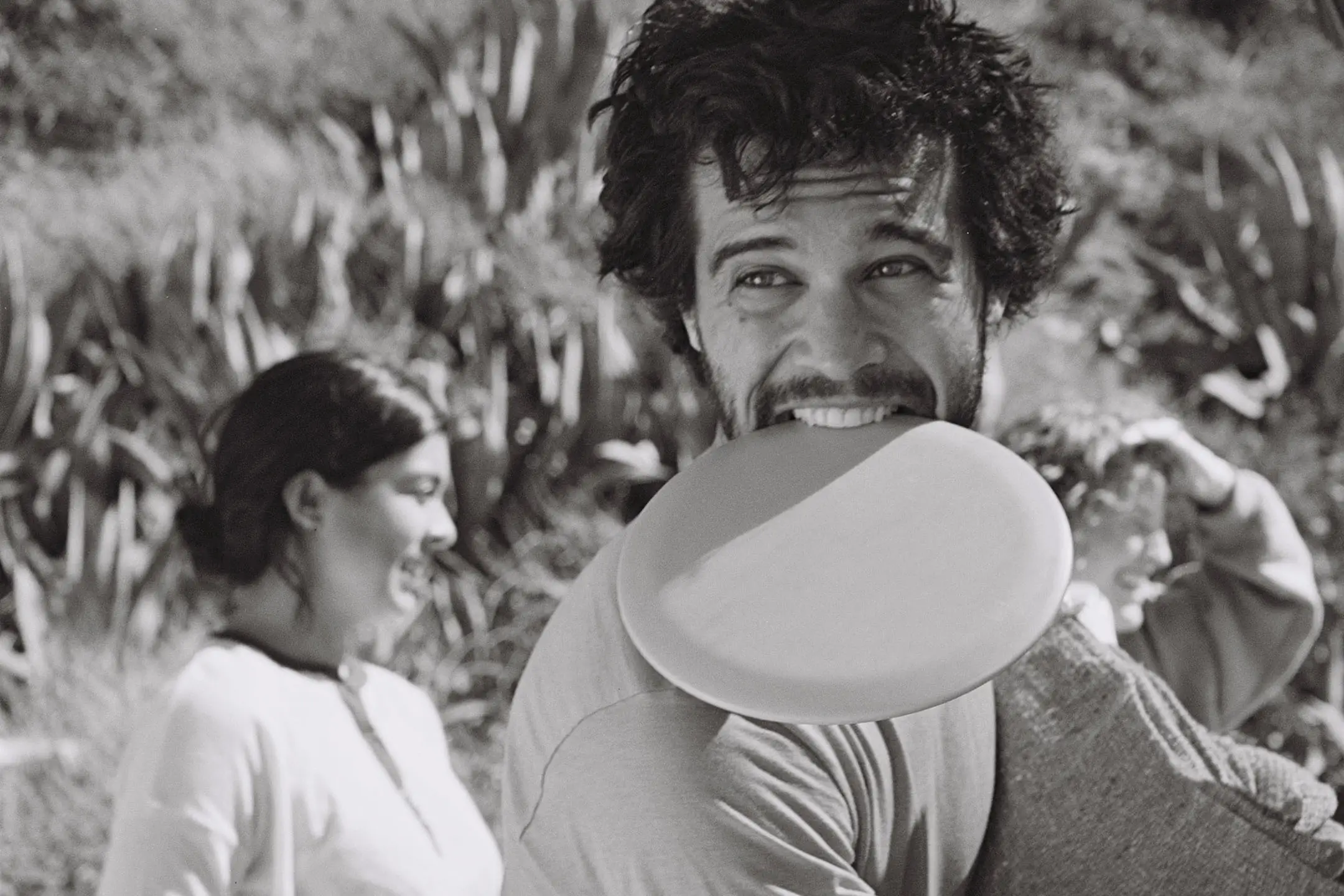 Man smiling while holding a frisbee in his mouth with two people laughing in the background in an outdoor setting.