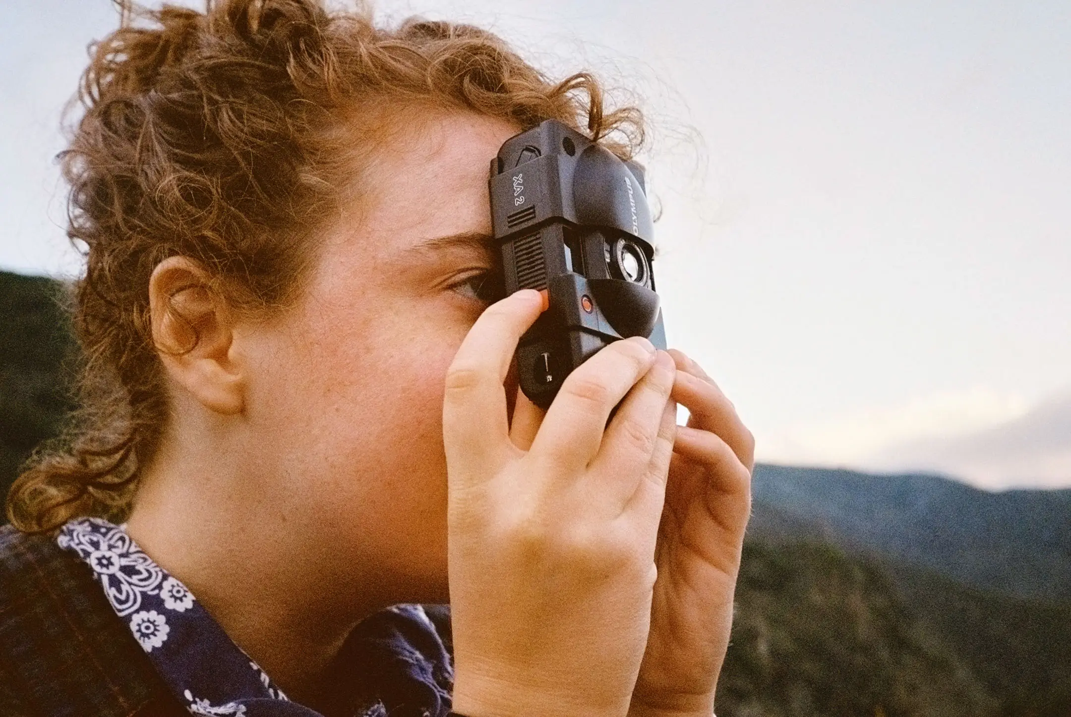 Person with curly hair using a vintage Olympus XA2 film camera outdoors near mountains.