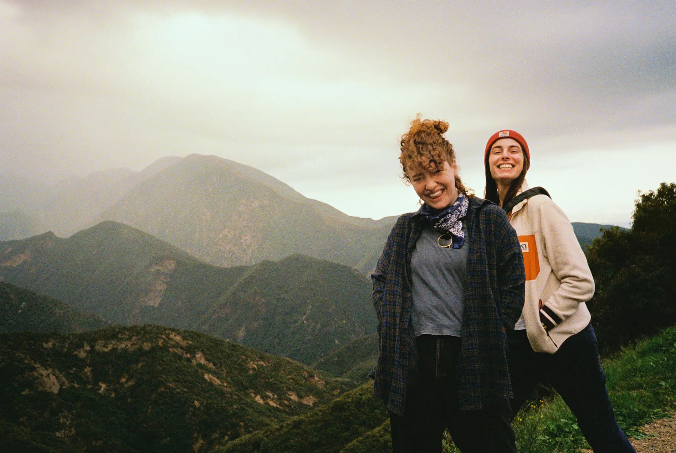 Two women smiling and posing on a grassy hillside with misty mountains in the background.