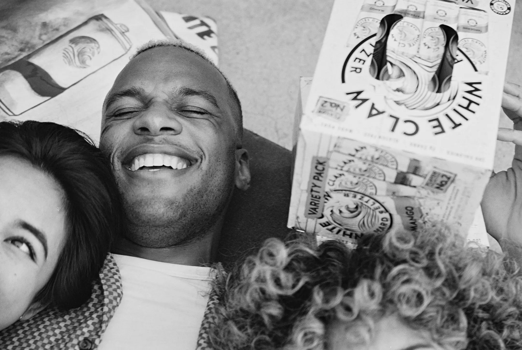 Black and white close-up of three friends lying down together, smiling and holding a White Claw variety pack.