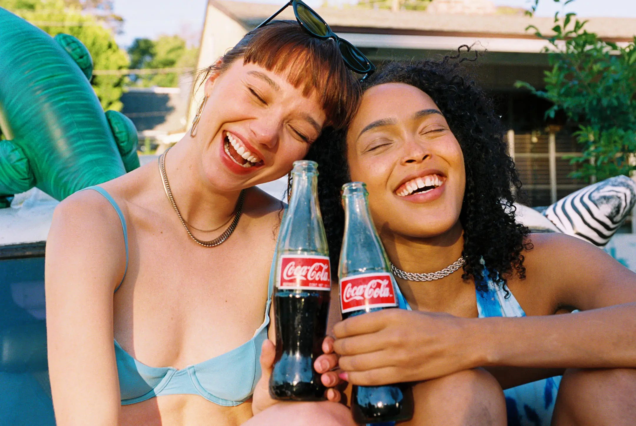 Two women smiling and holding glass Coca-Cola bottles while sitting outdoors on a sunny day.