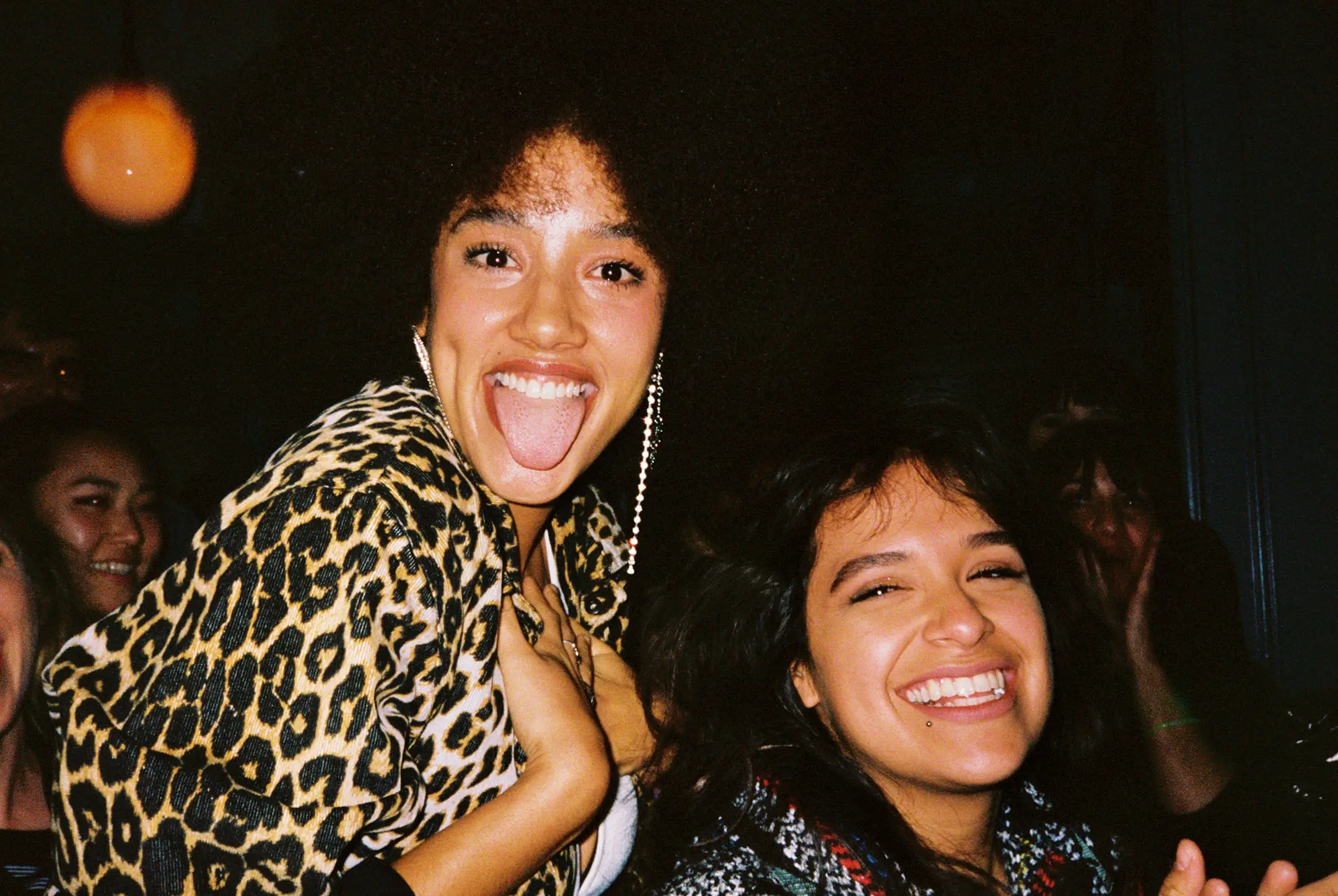 Two young women smiling and enjoying themselves at a party, one wearing a leopard print jacket and sticking out her tongue.