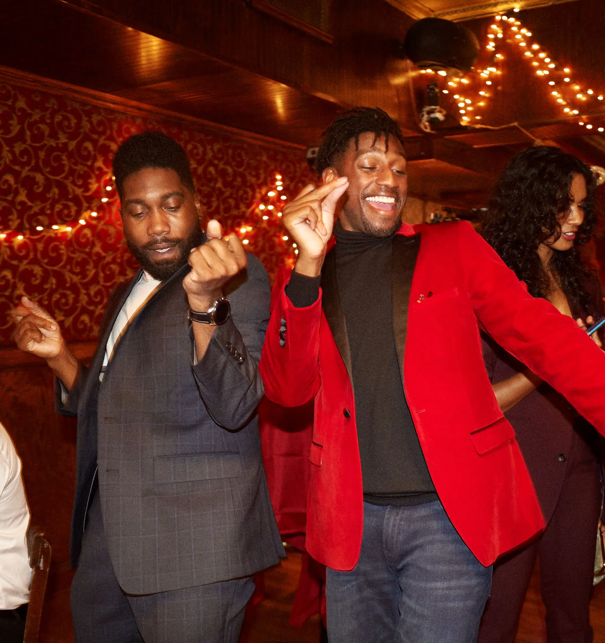 Two men dancing joyfully at a festive indoor party with string lights and red decorative wallpaper, a woman beside them looking at her phone.