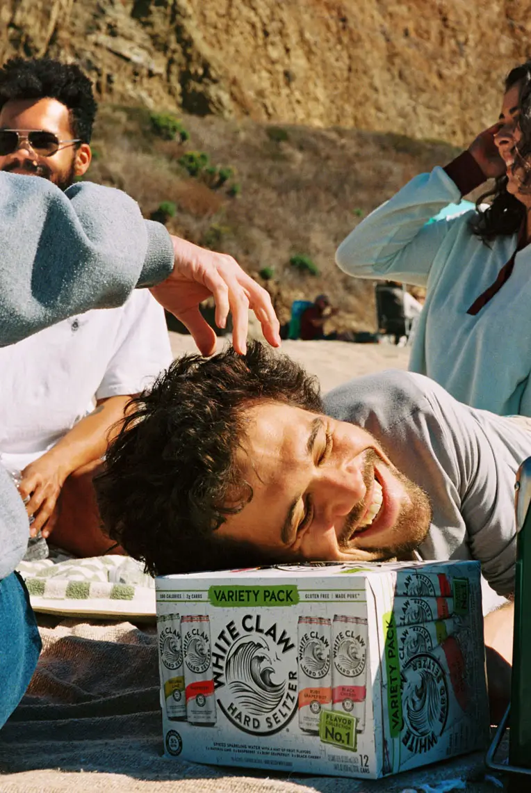 A man smiling with his head resting on a White Claw hard seltzer variety pack on a beach, surrounded by friends.