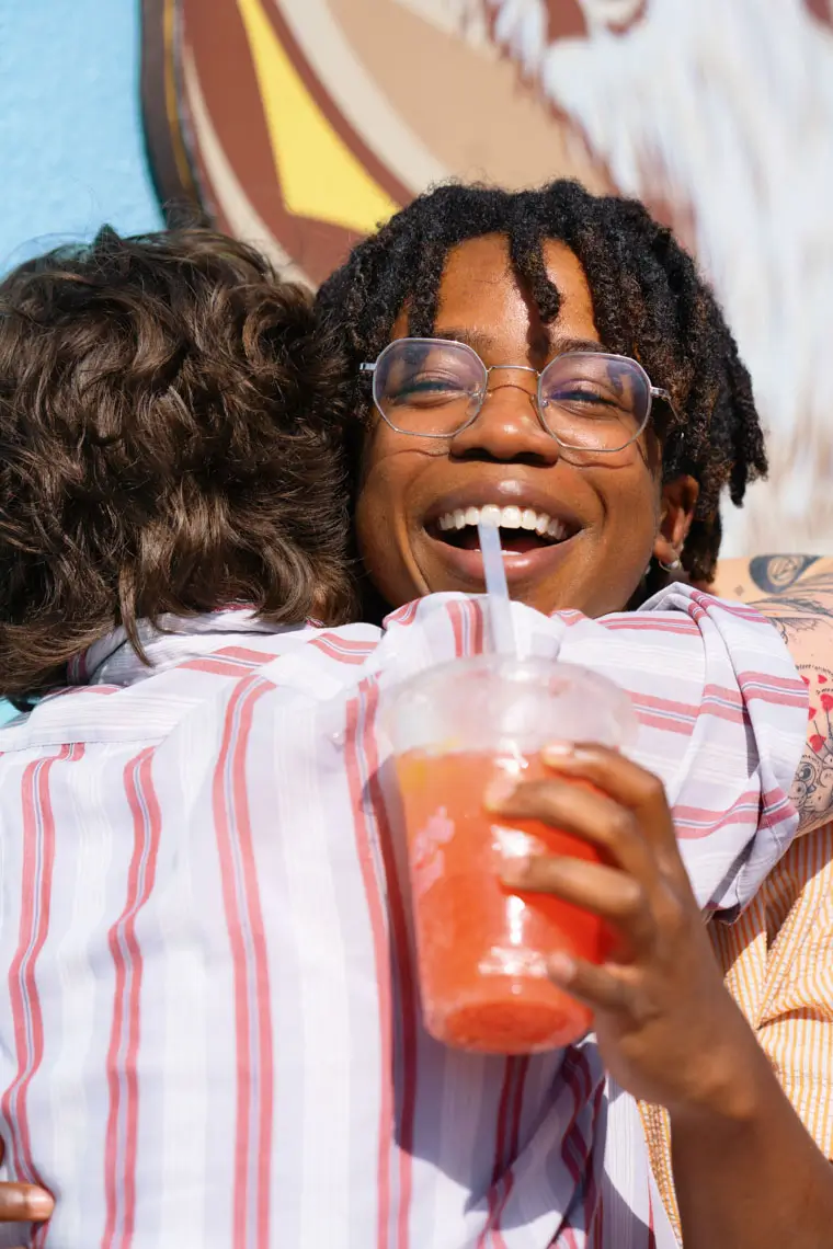 Two people embracing outdoors, one smiling and holding a red iced drink with a straw.