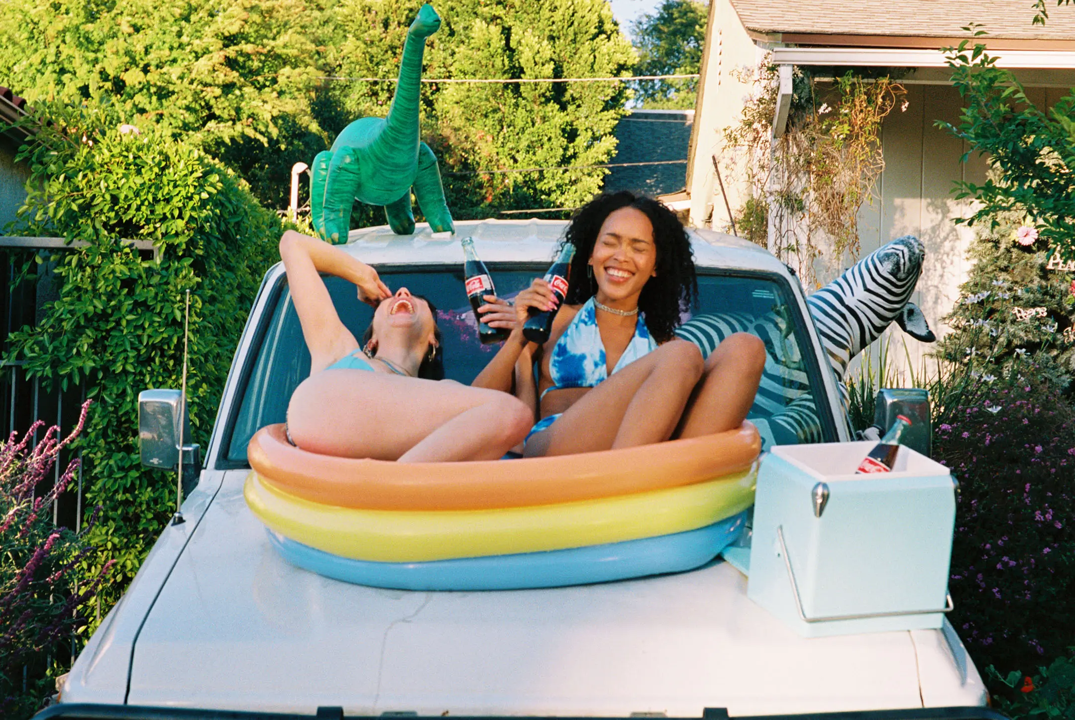 Two women in bikinis sitting on the hood of a white vehicle inside a small inflatable pool, holding glass bottles of Coca-Cola and laughing, with inflatable dinosaur and zebra on the vehicle.