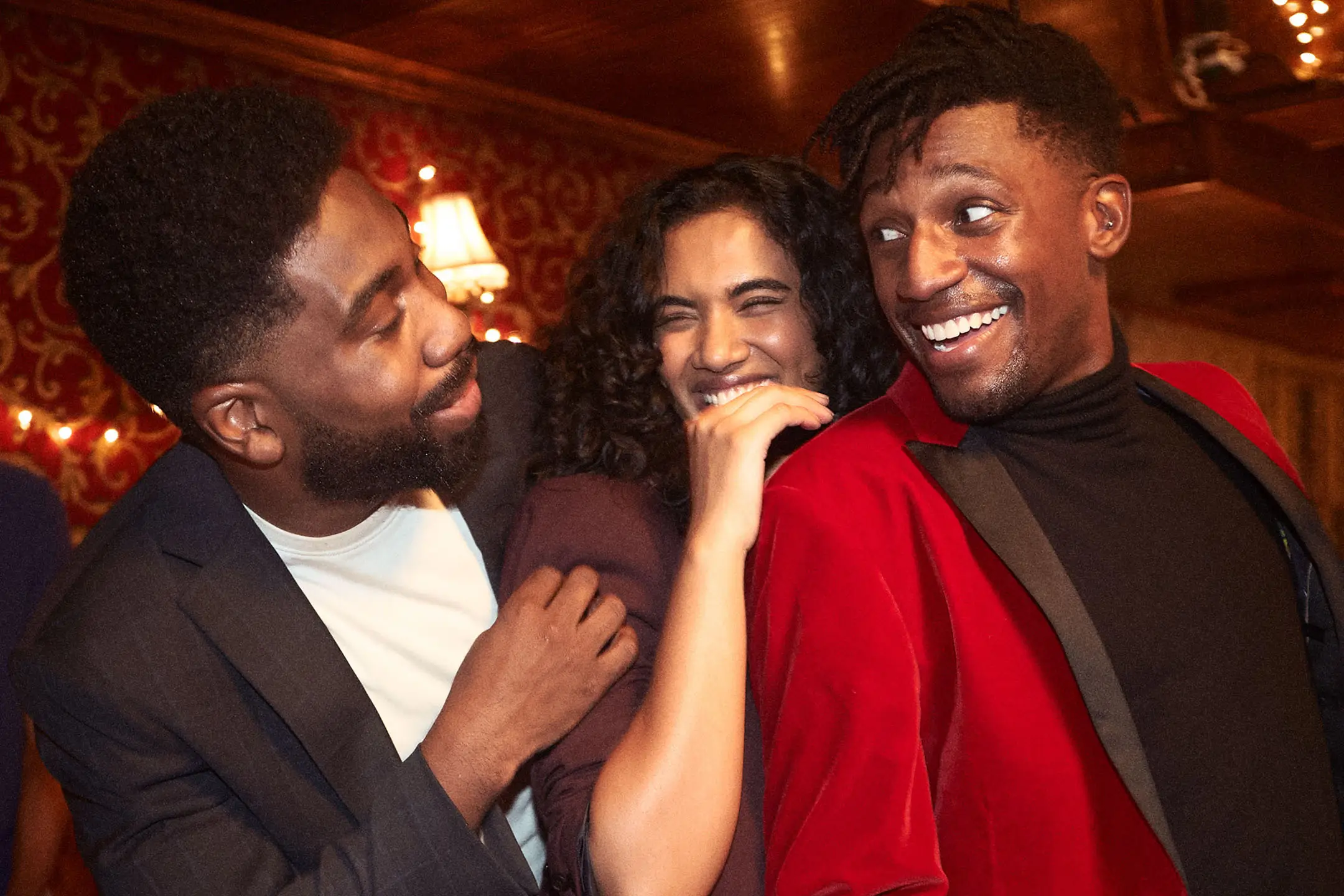 Three friends laughing and enjoying a festive indoor party, with one man wearing a red velvet jacket.
