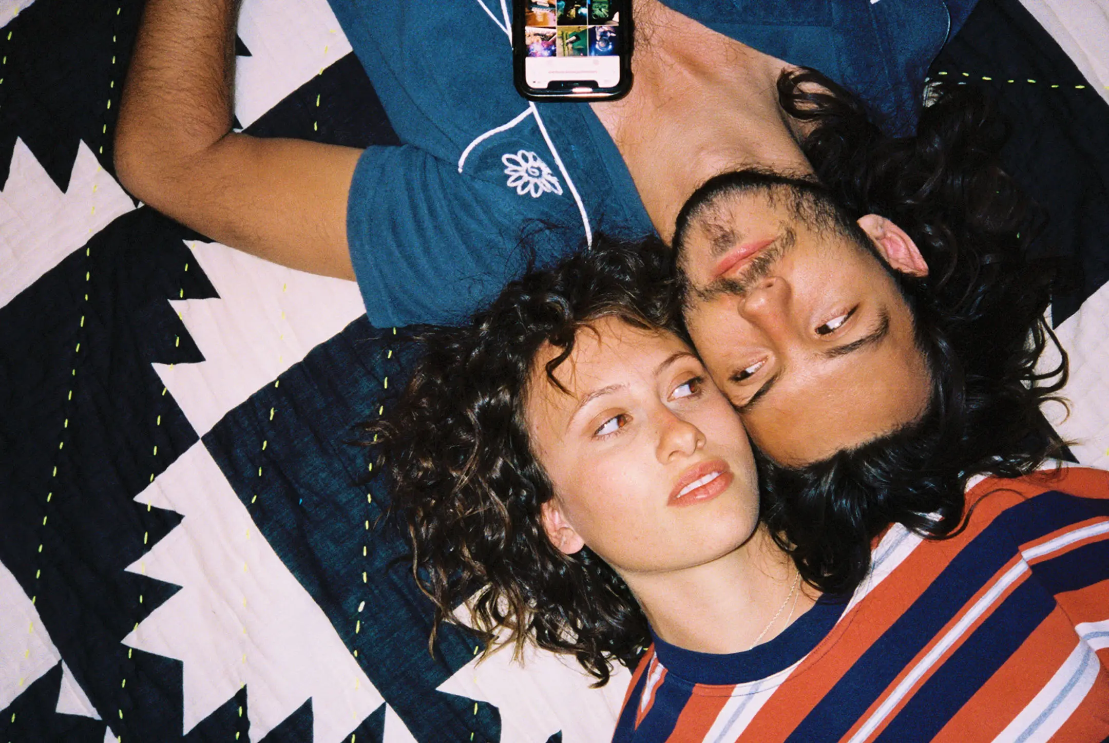 A couple lying on a patterned quilt; woman with curly hair in striped shirt and man with long hair and beard in blue shirt, looking at each other.