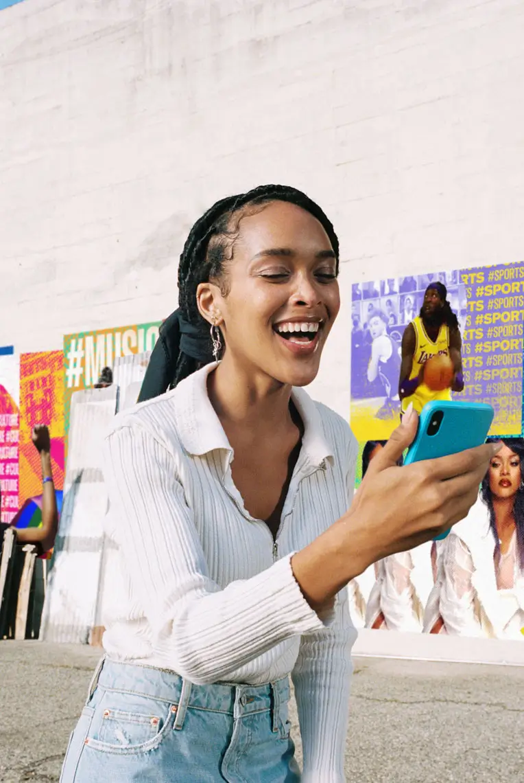 Smiling young woman with braided hair and hoop earrings looking at a blue smartphone outdoors near colorful posters.