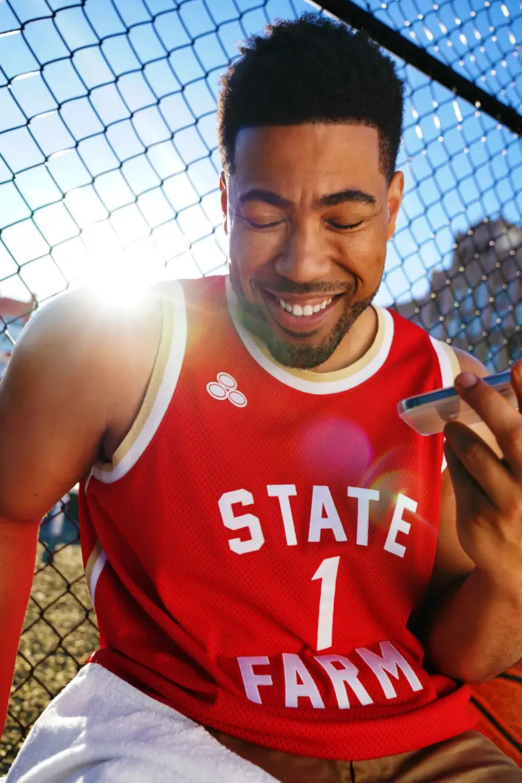 Smiling man in red basketball jersey with 'STATE FARM' and number 1, holding a phone near a chain-link fence with sunlight behind.