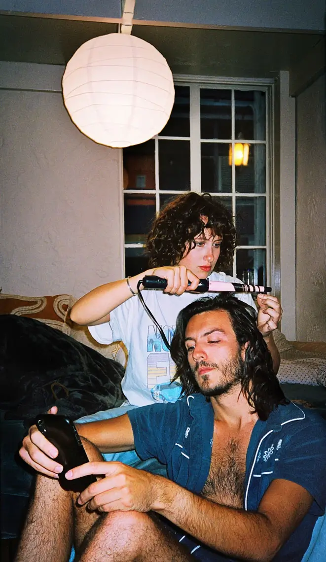 Woman with curly hair using a curling iron on a man's long hair as he looks at a phone, sitting on a couch under a round white hanging lamp.