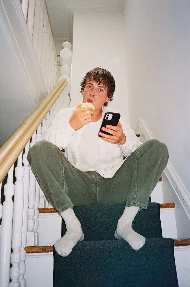 Young man sitting barefoot on carpeted stairs eating a sandwich and holding a smartphone.