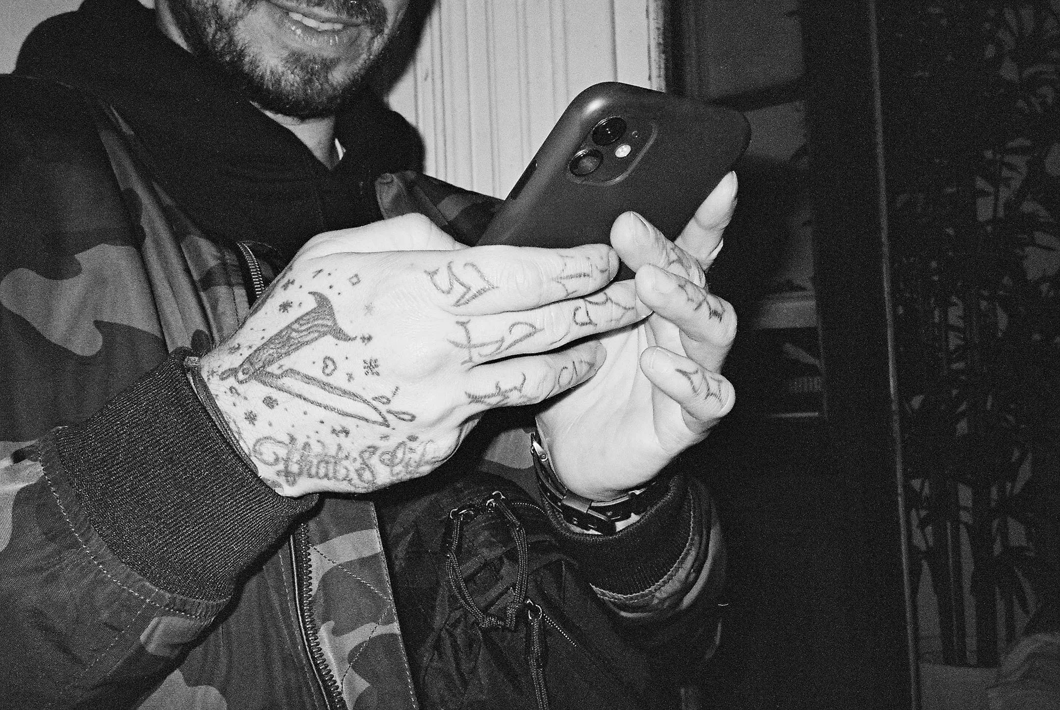 Person with tattooed hands holding and using a smartphone, wearing a camouflage jacket and a watch.