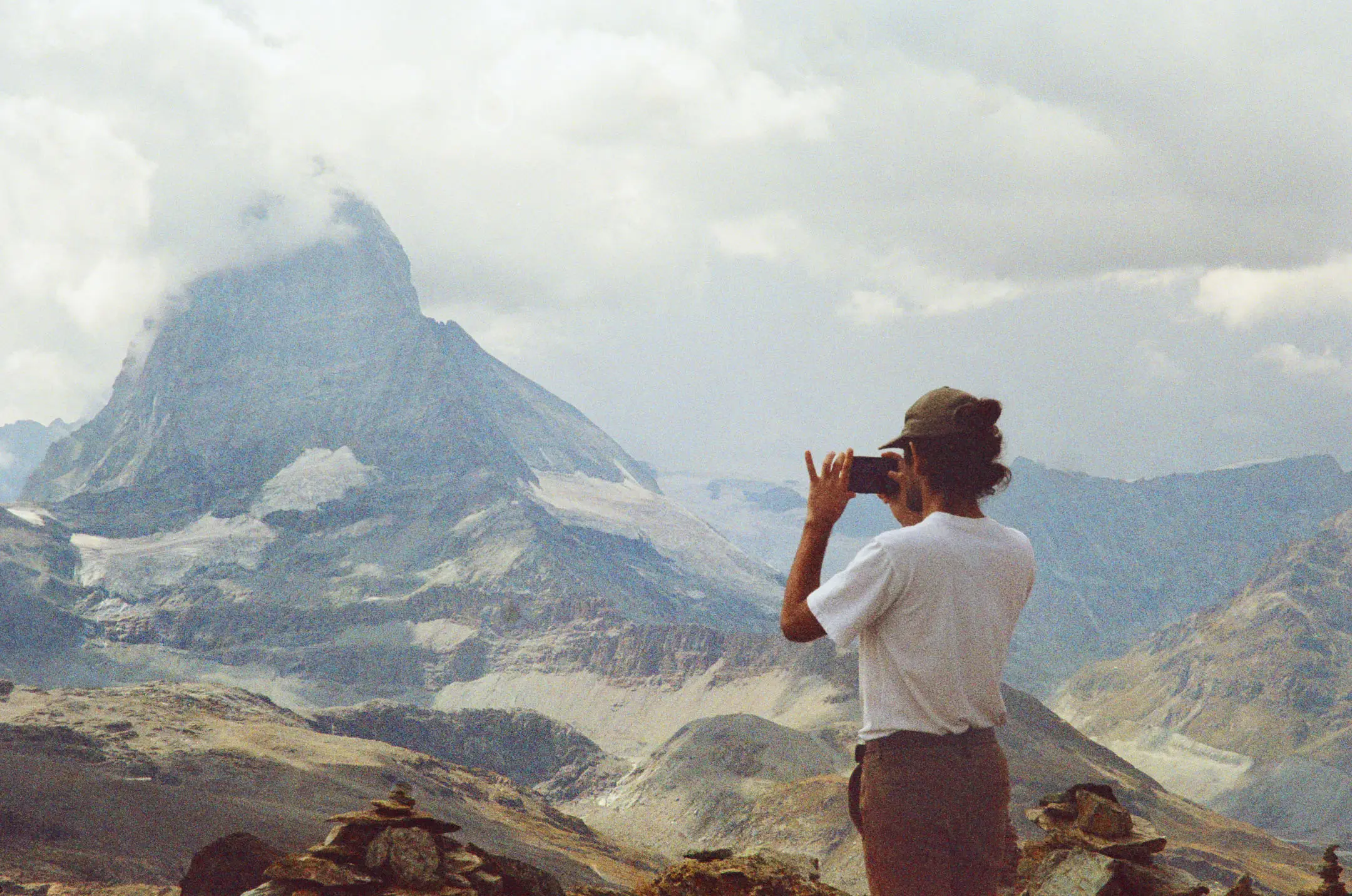 Person wearing a cap taking a photo of a towering mountain partially covered by clouds during a cloudy day.