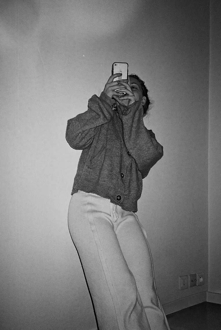 Person wearing a sweater and light pants taking a photo with a smartphone indoors against a plain wall.