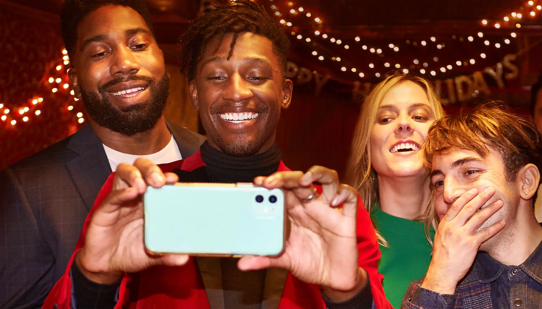 Four friends smiling and posing for a selfie at a holiday party with string lights in the background.