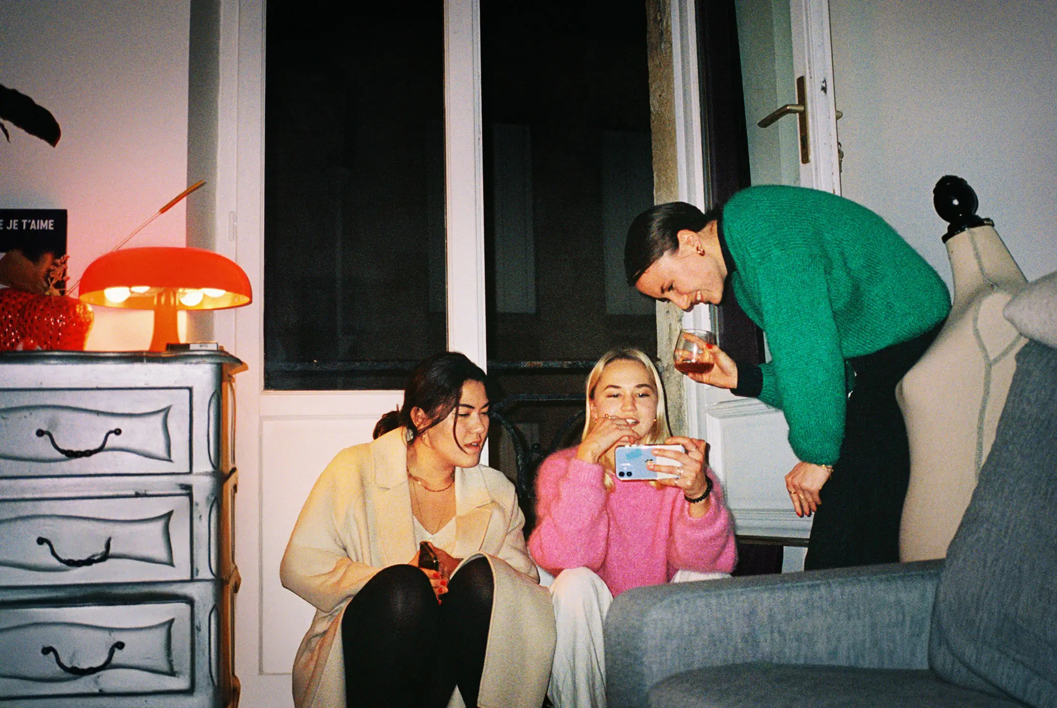 Three women indoors, two sitting on the floor looking at a phone, one standing leaning in smiling, holding a drink.